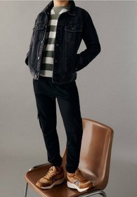 Svart denimjacka, gröngrå randig tröja, svarta byxor och bruna sneakers med blå accenter. Står på en brun stol.