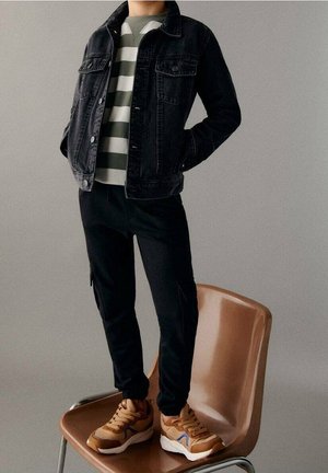 Svart denimjacka, gröngrå randig tröja, svarta byxor och bruna sneakers med blå accenter. Står på en brun stol.