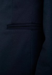 Gros plan d'une poche de veste en tissu bleu marine avec un passepoil horizontal et des détails de couture visibles.