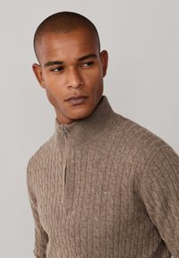 Lichtbruine kabelbreipullover met een opstaande kraag en een halve rits. Zachte textuur met subtiele logo-details op de borst.