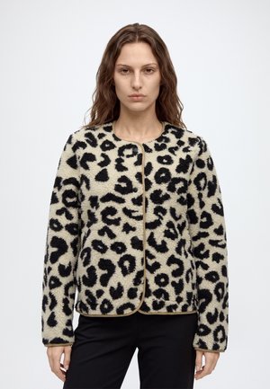 Femme aux longs cheveux ondulés porte une veste polaire beige et noire à imprimé léopard avec un col rond et un pantalon noir sur un fond blanc uni.