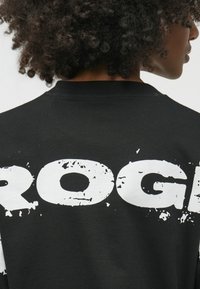 Melnas kokvilnas T-krekls ar lielu, nolietotu balto uzrakstu uz aizmugures. Apaļa kakla izgriezuma un īsas piedurknes pabeidz dizainu.