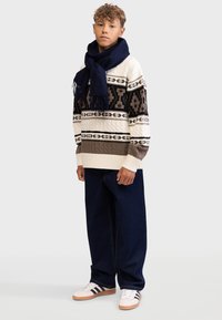 Striktrøje med geometriske mønstre i beige, creme og sort; parret med mørkeblå brede bukser og et mørkeblåt tørklæde.