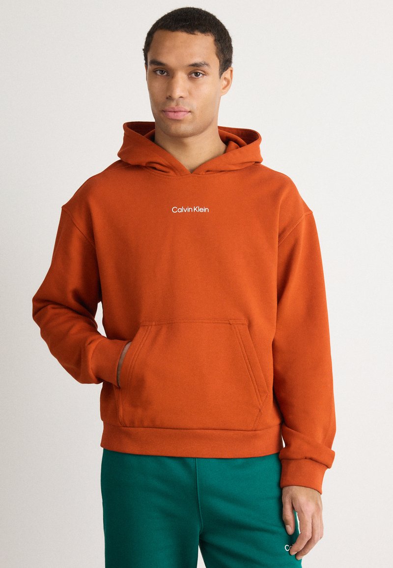 Felpa con cappuccio color ruggine, dotata di tasca a marsupio e piccolo logo bianco "Calvin Klein" sul petto. Tessuto morbido, vestibilità comoda, polsini a costine.