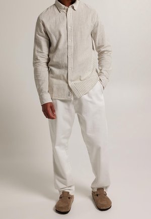 Mężczyzna ubrany w beżowo-białą koszulę w pionowe paski na guziki, białe luźne spodnie oraz brązowe zamszowe buty wsuwane, stojący na jednolitym tle.