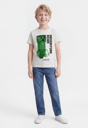 Smilende dreng iført en hvid Minecraft T-shirt med et grønt Creeper-grafik og teksten "Adventure Mode", blå jeans og hvide sneakers.
