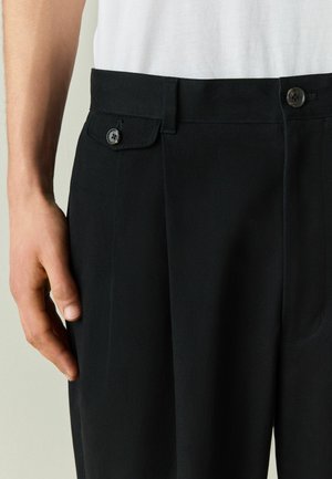 Mann trägt schwarze, maßgeschneiderte Hose mit geknöpfter seitlicher Taschenklappe und weißem Hemd, Hand ruht neben der Hose.