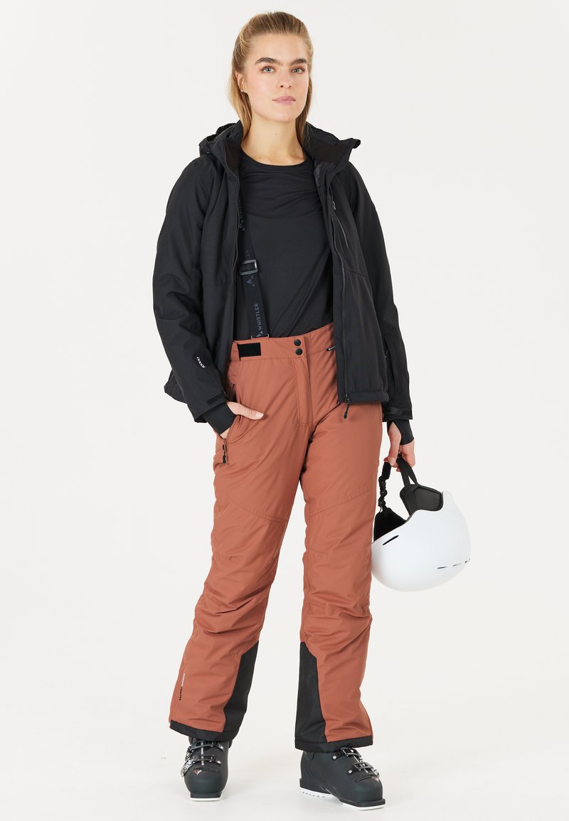 Skidkläder som inkluderar en svart isolerad jacka, långärmad svart topp, bruna skidbyxor med svarta accenter och en vit hjälm.