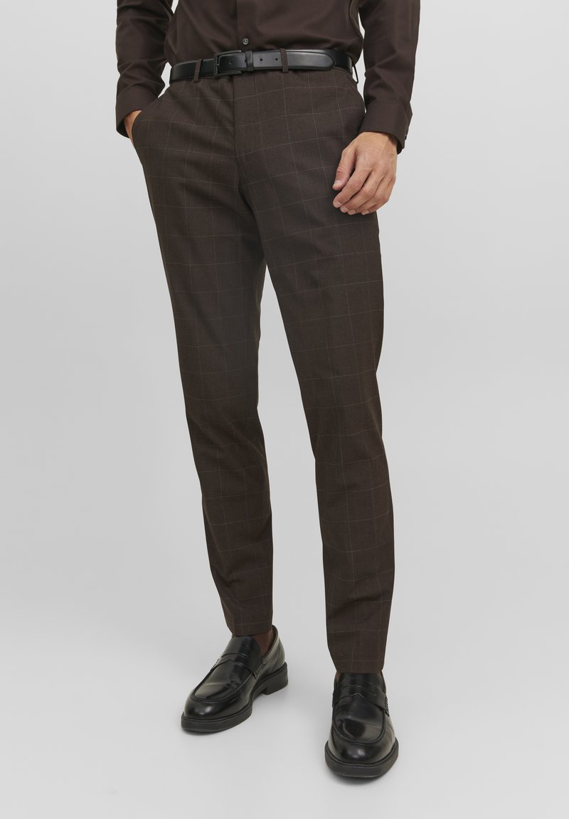 Pantaloni a quadri marroni con vestibilità su misura, caratterizzati da una texture liscia, passanti per la cintura e tasche laterali. Abbinati a mocassini neri.