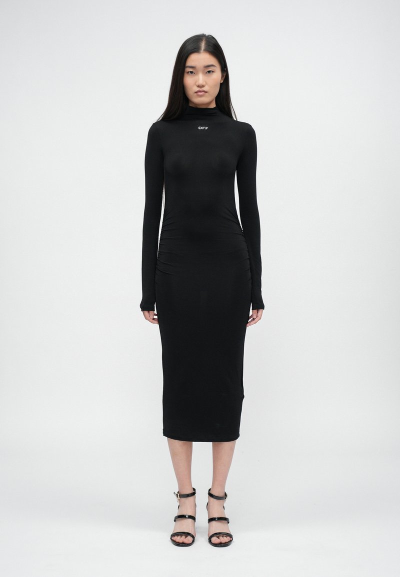 Čierne bodycon šaty s dlhým rukávom a vysokým golierom, so skladanými detailmi po stranách a priliehavým strihom. Nosia sa s čiernymi sandálmi na opätku.