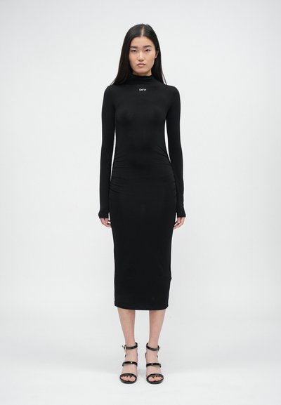 Czarna długa sukienka typu bodycon z wysokim kołnierzem, zebranymi detalami po bokach i dopasowanym krojem. Noszona z czarnymi sandałkami na obcasie.