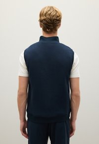 Gilet in pile blu navy con colletto alto, senza chiusure e dalla texture liscia. Indossato sopra una maglietta bianca. Design minimalista, senza decorazioni.