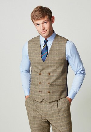 Ung mand iført en brun rutet vest og bukser med en lyseblå skjorte og blå stribet slips, stående med hænderne i lommerne.