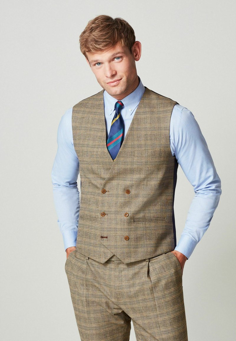 Mladý muž v hnedom kockovanom vestu a nohaviciach s svetlomodrou košeľou a modrou pruhovanou kravatou, stojí so založenými rukami v kapsách.