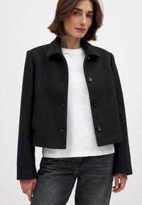 Schwarze kurz geschnittene Jacke aus weichem Stoff, mit einem Kragen, Knöpfen vorne und Seitentaschen. Trägt eine weiße Bluse und graue Jeans.
