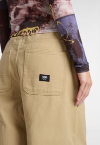 Pantalones cargo beige hechos de tela resistente, con dos bolsillos traseros y un parche de logo negro de Vans, parcialmente cubierto por una blusa con patrón.