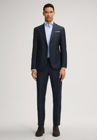 Abito blu navy con design aderente, giacca monopetto e pantaloni slim. Camicia da uomo azzurro chiaro con colletto. Stivaletti marroni alla caviglia.