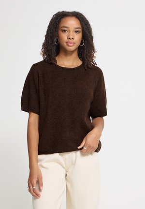 Kvinde med krøllet hår iført en mørkebrun kortærmet sweater og lyse beige bukser, stående mod en ensfarvet lys baggrund.