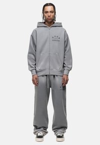 Felpa grigia con zip e pantaloni della tuta abbinati in tessuto morbido. La felpa presenta una tasca frontale; entrambi i pezzi hanno sottili accenti di testo stampato.