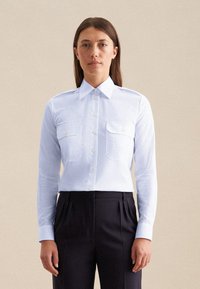 Lange mouw lichtblauw overhemd met twee borstzakken, knoopsluiting bij de kraag en gouden knopen, gecombineerd met donkere geplooide broek. Glad stoftextuur.
