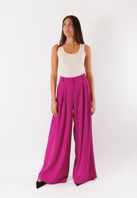 Pantaloni a vita alta, con pieghe in tessuto rosa vivace, taglio a gamba larga. Abbinati a un top senza maniche beige e tacchi a punta neri.