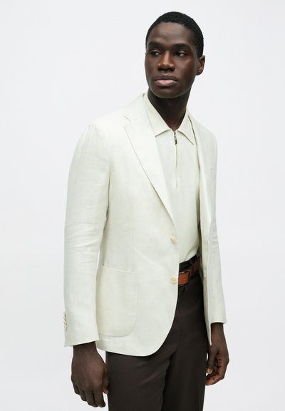 Homme portant un blazer crème sur une chemise crème zippée, avec un pantalon marron foncé et une ceinture marron, regardant légèrement vers sa gauche.