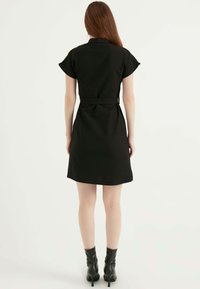 Schwarzes Kleid mit kurzen Ärmelchen, taillierter Taille und einem Gürtel-Detail. Glatte Textur, knielang, kombiniert mit schwarzen Absatzstiefeln.