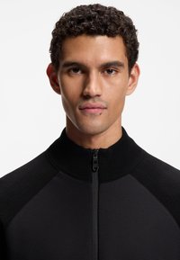 Μαύρο μπουφάν με φερμουάρ, με υφή υφάσματος, ψηλό γιακά και λείο διακοσμητικό στα σημεία των ώμων και των μανικιών. Απλός και λειτουργικός σχεδιασμός.