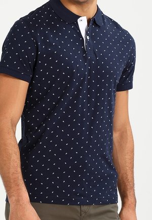 Mann trägt ein dunkelblaues Poloshirt mit kleinen weißen Paisley-Mustern und beigen Hosen, steht vor einem schlichten Hintergrund.