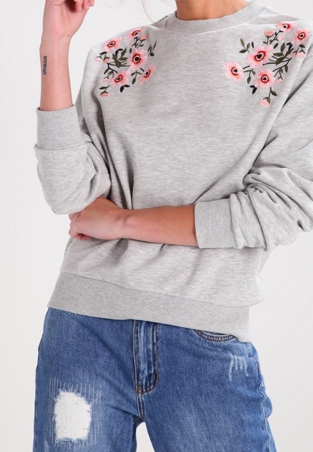 Felpa grigia con ricami floreali in rosa e verde sulle spalle. Polsini e bordo a costine. Indossata con jeans blu strappati.