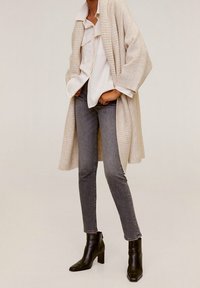 Personne portant un long cardigan crème, une chemise blanche à boutons, un jean slim gris et des bottines noires à talons, se tenant les mains dans les poches.