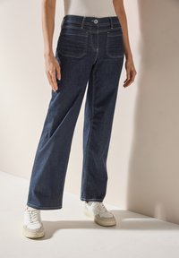 Pantalons en denim à jambe large de couleur bleu foncé avec des coutures contrastantes, fermeture à bouton à l'avant et poches latérales, associés à des baskets blanches.