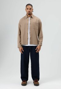 Homme debout portant une chemise beige à boutons par-dessus un t-shirt blanc, un pantalon large foncé et des bottes marron à lacets sur un fond uni.