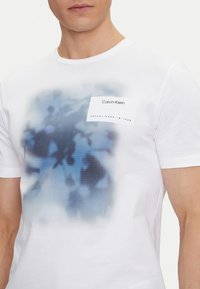 Camiseta de algodón blanca con un estampado abstracto azul en el frente y una etiqueta de tela con "Calvin Klein" y el año "1968".