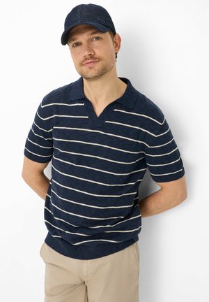Mann trägt ein navyblau gestreiftes kurzärmeliges Poloshirt, beige Hose und navyblaue Kappe, steht mit den Händen hinter dem Rücken vor weißem Hintergrund.