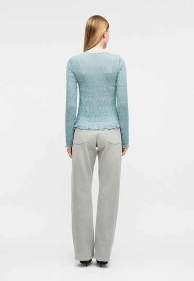 Γυναίκα με μακριά ξανθά μαλλιά που φοράει μπλε ανοιχτόχρωμο μακρυμάνικο τοπ με υφή και φαρδιά γκρι ανοιχτό παντελόνια, όρθια με την πλάτη προς το μέρος μας.