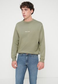 Φούτερ σε ελιά πράσινο με στρογγυλή λαιμόκοψη, ριμπ μανσέτες και κεντημένο κείμενο στο στήθος, σε συνδυασμό με ανοιχτό μπλε τζιν παντελόνι.