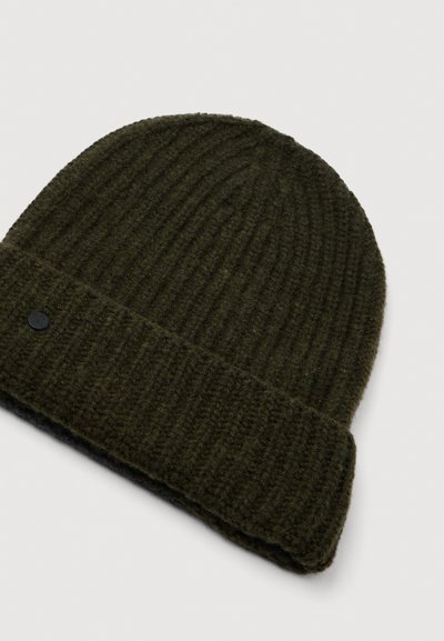 Adīta tumši zaļa vilnas beanie cepure, ar ribotu tekstūru, salocītu maliņu un nelielu melnu logotipa akcentu uz sāna.
