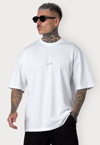 White cotton t-shirt with a relaxed fit and short sleeves, featuring a small printed logo on the chest. Black sunglasses, tattooed arm visible.