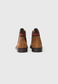 Bruine suède enkelboots met zwarte elastische zijpanelen en rode logo-accenten bij de hak. Platte rubberen zool, gladde afwerking.