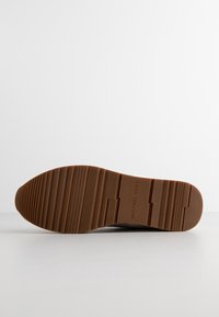 Semelle de chaussure marron avec motif rainuré et inscription "MICHAEL KORS" embossée au centre sur fond blanc.