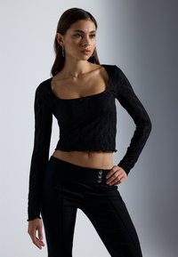 Schwarzes Spitzenoberteil mit langen Ärmeln und quadratischem Ausschnitt sowie gewelltem Saum, kombiniert mit hochgeschnittenen schwarzen Hosen mit silbernen Knopfakzenten.
