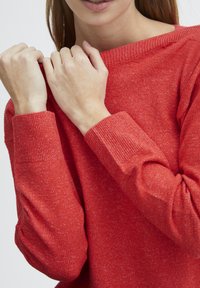 Pull en tricot rouge avec une surface texturée, un col bateau et des poignets côtelés. Le tissu semble doux et légèrement chiné.