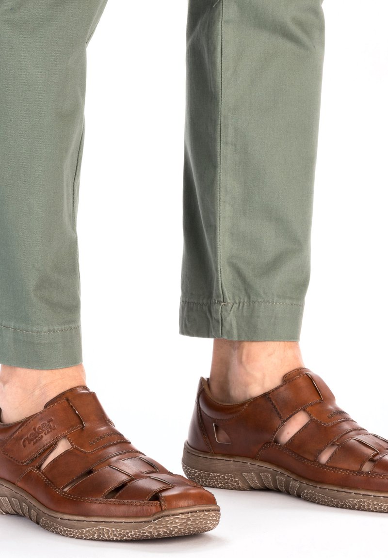 Braune Leder-Sandalen mit ausgeschnittenen Designs, strukturierten Sohlen und verstellbaren Riemen, kombiniert mit hellgrünen Knöchellangen Hosen.