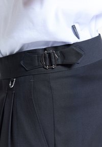 Chemise blanche rentrée dans un pantalon noir avec une petite boucle métallique décorative et un détail de sangle près de la ceinture.