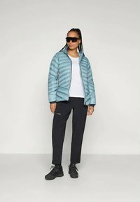 Žena u crnim naočalama, svijetloplavoj jakni s čvrstim punjenjem, bijeloj majici, crnim hlačama i crnim tenisicama stoji naslonjena na ravnu pozadinu.