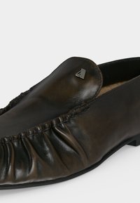 Μαύρο δερμάτινο παπούτσι τύπου slip-on με πιετά λεπτομέρειες κατά μήκος της πλευράς και μικρό μεταλλικό λογότυπο στο μπροστινό πάνω μέρος.