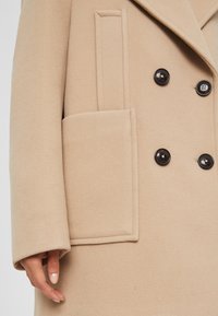 Abrigo beige de doble botonadura con un acabado texturizado similar a la lana, que presenta grandes botones negros y un bolsillo lateral con un detalle de costura en el borde.