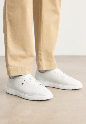 White leather sneakers with a smooth finish, featuring round toe, lace-up design, and subtle logo detail on the side, paired with beige pants.