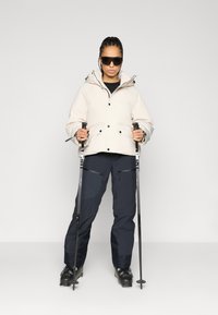 Σακάκι σκι σε ανοιχτό μπεζ με κουκούλα, μαύρα κουμπιά και τσέπες. Συνδυασμένο με σκούρα μπλε παντελόνια σκι και μαύρες μπότες σκι, κρατώντας μπαστούνια σκι.